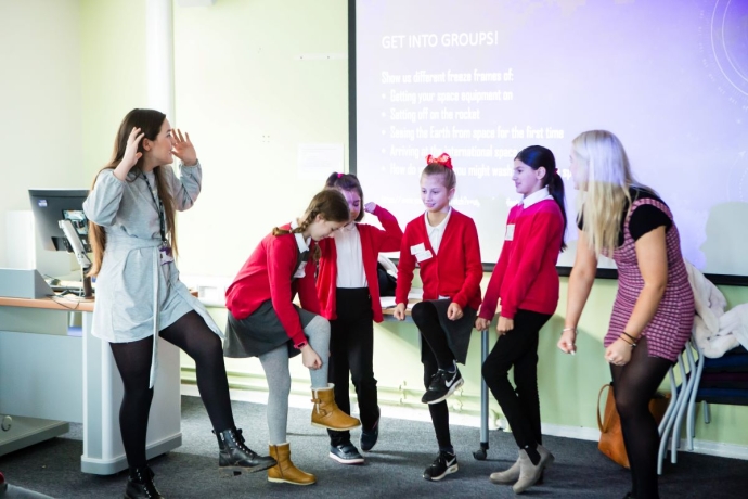 In a classroom, five children in red sweaters and one adult participate in an activity involving lifting one leg. A projector screen in the background displays group activity instructions titled 'GET INTO GROUPS!' with prompts related to space exploration.